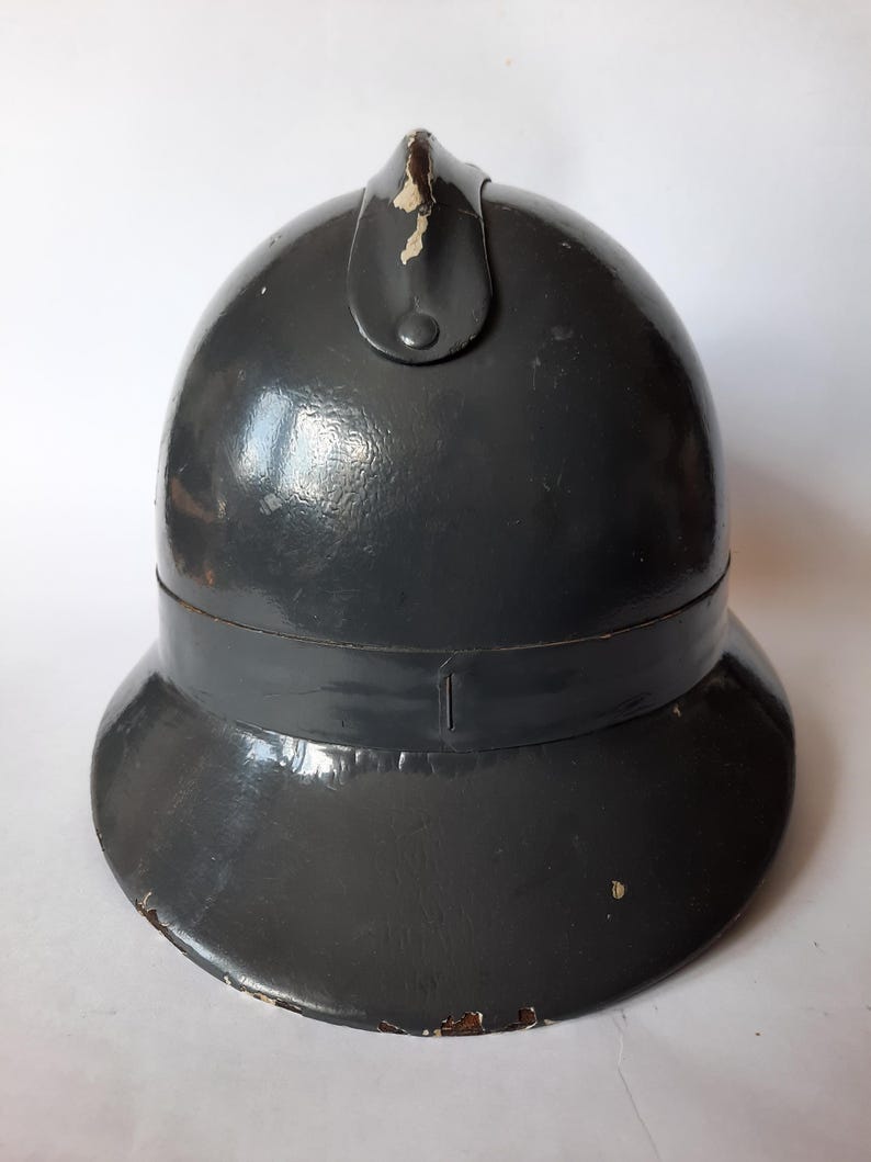 K&ouml;nnte beinhalten: Ein dunkelgrauer, gl&auml;nzender, abgerundeter Helm mit einem zentralen vertikalen Grat an der Oberseite. Der Helm hat eine breite, ausgestellte Krempe und weist sichtbare Gebrauchsspuren auf, darunter Farbabsplitterungen am Kamm und am Rand.