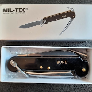 以下が含まれることがあります： 銀色の刃と先端にフックが付いた黒い折りたたみナイフ。ナイフのハンドルには「BUND」と印刷されています。ナイフは、「MIL-TEC® Tactical & Outdoor Products」と印刷された白い箱に入っています。