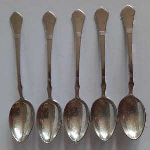 May include: Five silver spoons arranged on a white surface. Each spoon has an oval bowl and a long handle with a decorative, geometric design near the top. The spoons are reflective, showing the surrounding environment.