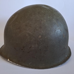 Può includere: Un elmetto militare a forma di cupola di colore verde oliva con una calotta arrotondata e una tesa leggermente svasata. La superficie dell'elmetto mostra segni di usura con graffi e scalfitture visibili, rifinito con un bordo in metallo.
