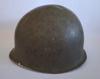 May include: A dome-shaped olive drab military helmet with a smooth, rounded crown and a slightly flared brim. The surface of the helmet shows signs of wear with visible scuffs, scratches, and a matte finish, featuring a metal rim edge.