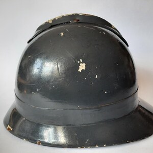 K&ouml;nnte beinhalten: Ein dunkelgrauer Metallhelm mit einer abgerundeten Kuppel und einer breiten Krempe. Die Oberfl&auml;che weist Gebrauchsspuren mit sichtbaren Kratzern und kleinen Stellen auf, an denen die Farbe abgebl&auml;ttert ist und eine hellere Farbe freigibt.