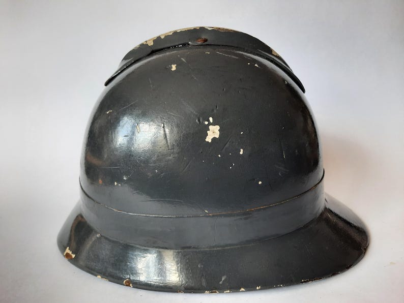 K&ouml;nnte beinhalten: Ein dunkelgrauer Metallhelm mit einer abgerundeten Kuppel und einer breiten Krempe. Die Oberfl&auml;che weist Gebrauchsspuren mit sichtbaren Kratzern und kleinen Stellen auf, an denen die Farbe abgebl&auml;ttert ist und eine hellere Farbe freigibt.