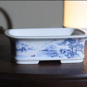 Könnte beinhalten: Ein rechteckiger weißer Keramik-Pflanztopf mit blauem Landschaftsdesign. Der Pflanztopf zeigt eine detaillierte Illustration von Bergen, Bäumen und Figuren. Das Design umschließt die Seiten des Pflanztopfs. Der Pflanztopf hat eine klassische, elegante Ästhetik.