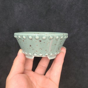 Puede incluir: Una maceta pequeña de cerámica azul claro con un esmalte moteado y un borde elevado. La maceta tiene tres pequeños pies y es perfecta para plantas pequeñas.