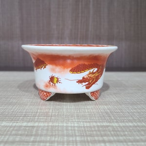 Può includere: Un vaso di ceramica bianca con tre gambe e un design di drago rosso e arancione. Il vaso ha un design di drago rosso e arancione con un globo giallo.