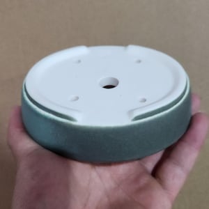 May include: A round, ceramic plant pot saucer with a white interior and a teal exterior. The saucer has a central drainage hole and several smaller holes. The pot is held in a hand.