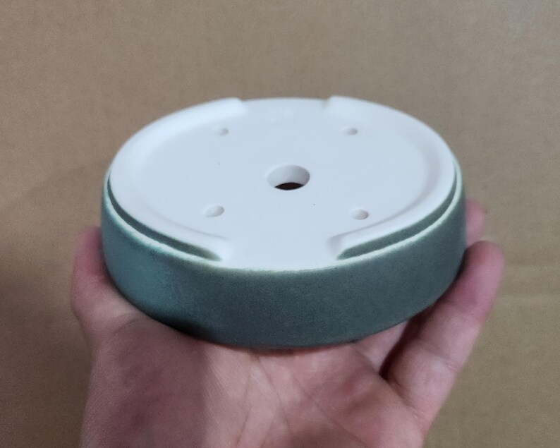May include: A round, ceramic plant pot saucer with a white interior and a teal exterior. The saucer has a central drainage hole and several smaller holes. The pot is held in a hand.