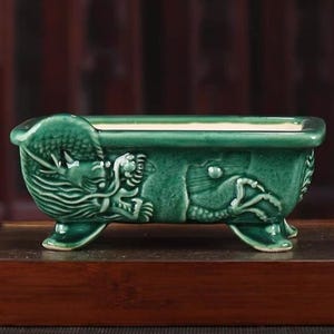 Könnte beinhalten: Ein rechteckiger, smaragdgrüner Keramik-Pflanztopf mit einem Drachenmotiv. Der Pflanztopf wird von drei kleinen Füßen getragen und weist detaillierte Reliefschnitzereien eines Drachen auf. Der Pflanztopf ist etwa 20 cm lang.