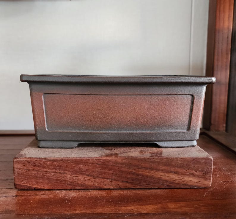 K&ouml;nnte beinhalten: Ein rechteckiger Bonsai-Topf mit dunkelbrauner, strukturierter Oberfl&auml;che. Der Topf steht auf einem Holzsockel mit r&ouml;tlich-braunem Farbton. Das Design des Topfes weist eine leicht erh&ouml;hte, rechteckige Platte auf der Vorderseite auf.