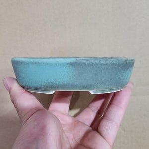 May include: A small, oval-shaped bonsai pot with a teal glaze. The ceramic pot has a low profile and is held in a hand. The pot has a textured surface and is designed for planting small trees or plants.