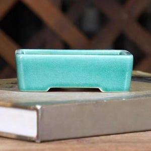 May include: A small, rectangular, teal-colored ceramic bonsai pot. The pot has a slightly raised lip around the top edge and a small lip on the bottom edge.