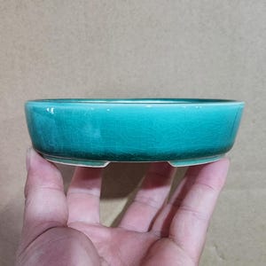 May include: A small, turquoise ceramic bonsai pot with a crackled glaze. The round, shallow dish has a low profile and three small feet. The pot is held in a hand against a neutral background.
