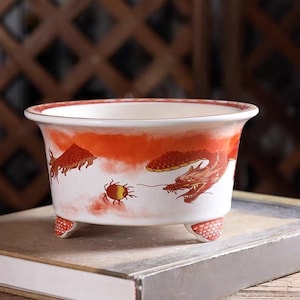 May include: A white ceramic planter with a red and orange dragon and fish design. The planter has three small feet and a wide rim. The design is in the style of traditional Asian art.