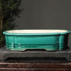 Puede incluir: Una maceta rectangular de cerámica color verde azulado, con interior crema. La maceta tiene una superficie texturizada y se apoya sobre un soporte de madera oscuro y ornamentado. El diseño de la maceta incluye crestas horizontales y bordes decorativos.