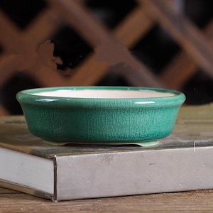 May include: A small, oval teal ceramic bonsai pot with a crackle glaze and a white interior. The pot sits on a book with a grey cover. The pot is designed for small plants. The pot's dimensions are in centimetres.