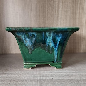 May include: A square, ceramic planter with a glossy, emerald green glaze. The glaze has a dripped effect with streaks of turquoise and blue. The planter has a rectangular base with four small feet.