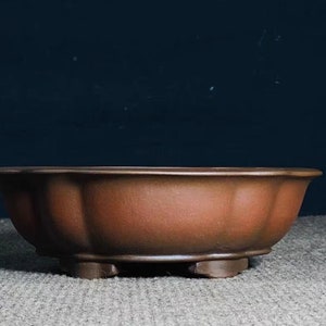 Puede incluir: Una maceta de bonsái de cerámica marrón con borde festoneado y tres pequeños pies. La maceta tiene un acabado mate y una forma ancha y poco profunda, adecuada para cultivar árboles o plantas pequeñas. El fondo es azul oscuro.