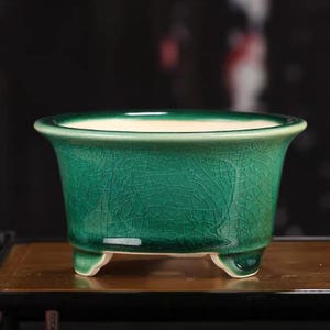 May include: A ceramic bonsai pot with a vibrant emerald green glaze featuring a distinct crackle pattern. The vessel has a wide, flared rim, a smooth cream-coloured interior, and is supported by three small, sturdy feet at the base.