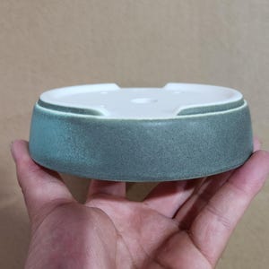May include: A round, ceramic ashtray with a white interior and a teal-colored exterior. The ashtray has a low profile and a slightly textured surface. The design includes a small notch on the side. The ashtray is held in a hand.