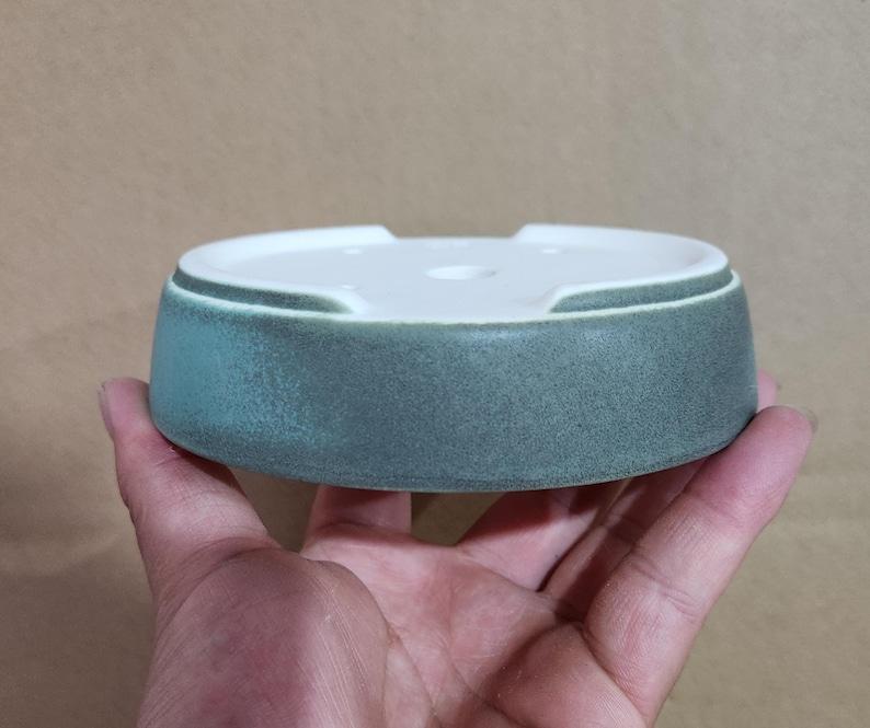 May include: A round, ceramic ashtray with a white interior and a teal-colored exterior. The ashtray has a low profile and a slightly textured surface. The design includes a small notch on the side. The ashtray is held in a hand.