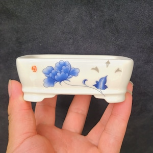 以下が含まれることがあります： 青とグレーの花柄が描かれた白い陶器製のプランター。プランターは3本の脚があり、長方形の形をしています。