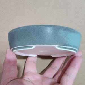 May include: A small, round, ceramic planter with a matte teal exterior and a pale pink interior. The planter has a low profile and a simple, modern design, suitable for small plants or succulents.