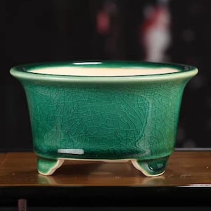 Op de afbeelding: Een kleine, groene keramische plantenbak met een craquelé-afwerking. De plantenbak heeft een brede, licht uitlopende rand en drie kleine voetjes. De binnenkant is lichtcrème van kleur.