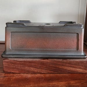 K&ouml;nnte beinhalten: Ein rechteckiger Bonsai-Topf mit dunklem, metallischem Finish, auf einem Holzsockel. Der Topf hat eine eingelassene Platte und eine leicht strukturierte Oberfl&auml;che. Der Holzsockel hat eine satte, rotbraune Farbe.