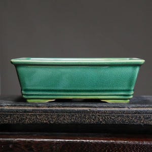 May include: A rectangular green ceramic bonsai pot with a textured glaze. The pot has a slightly raised rim and four small feet.