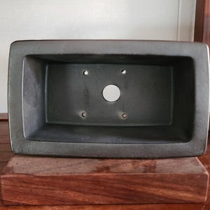 K&ouml;nnte beinhalten: Ein dunkelgrauer, rechteckiger Bonsai-Topf mit abgerundeten Ecken. Der Topf hat ein gro&szlig;es zentrales Abzugsloch und mehrere kleinere L&ouml;cher. Er steht auf einem dunklen Holzsockel und zeigt ein minimalistisches Design.