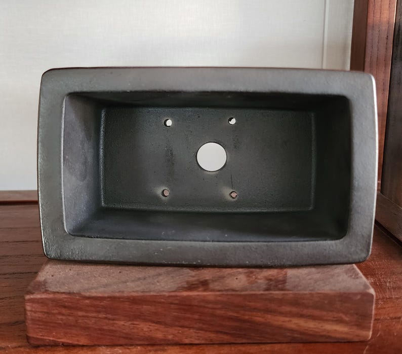 K&ouml;nnte beinhalten: Ein dunkelgrauer, rechteckiger Bonsai-Topf mit abgerundeten Ecken. Der Topf hat ein gro&szlig;es zentrales Abzugsloch und mehrere kleinere L&ouml;cher. Er steht auf einem dunklen Holzsockel und zeigt ein minimalistisches Design.