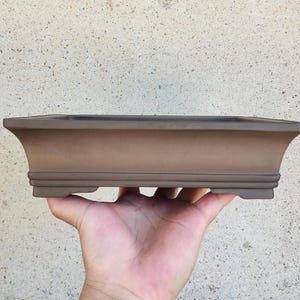 Könnte beinhalten: Ein brauner, rechteckiger Bonsai-Topf mit ausgestelltem Design. Der Topf hat eine breite, flache Form mit einer leicht strukturierten Oberfläche. Der Topf wird in einer Hand vor einem neutralen Hintergrund gehalten.
