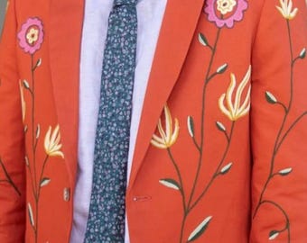 以下が含まれることがあります： ピンク、黄色、緑の刺繍の花柄があしらわれた鮮やかなオレンジ色のブレザー。ブレザーの下には、淡いピンクの襟付きシャツと、小さな白い花柄がプリントされたダークグリーンのネクタイを着用しています。明るい色のボタンが2つ見えます。