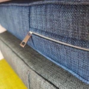 Puede incluir: Primer plano de un detalle de cremallera en una pila de tres cojines. El coj&iacute;n superior es de tela azul oscuro, el coj&iacute;n del medio es de tela gris oscuro y el coj&iacute;n inferior es de tela amarilla brillante.