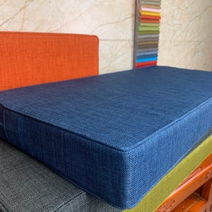 May include: A stack of four rectangular cushions in different colors. The top cushion is a deep blue, the second is a dark gray, the third is a light green, and the bottom is a dark brown wood.