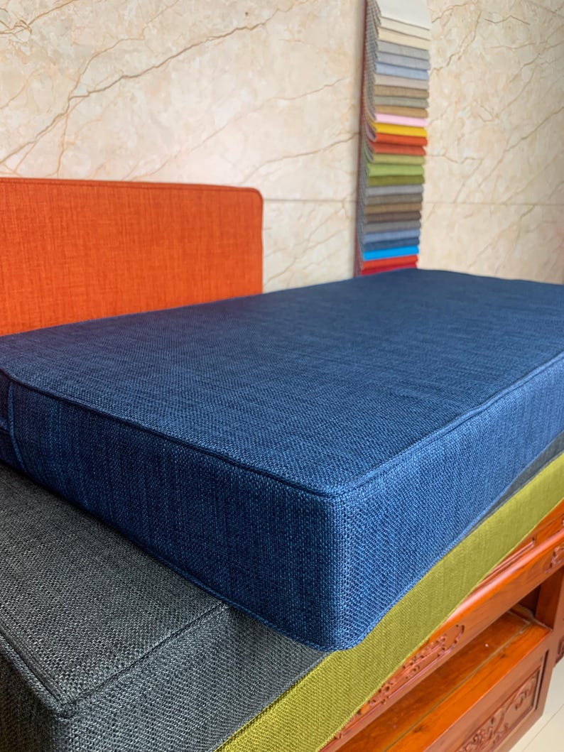 May include: A stack of four rectangular cushions in different colors. The top cushion is a deep blue, the second is a dark gray, the third is a light green, and the bottom is a dark brown wood.