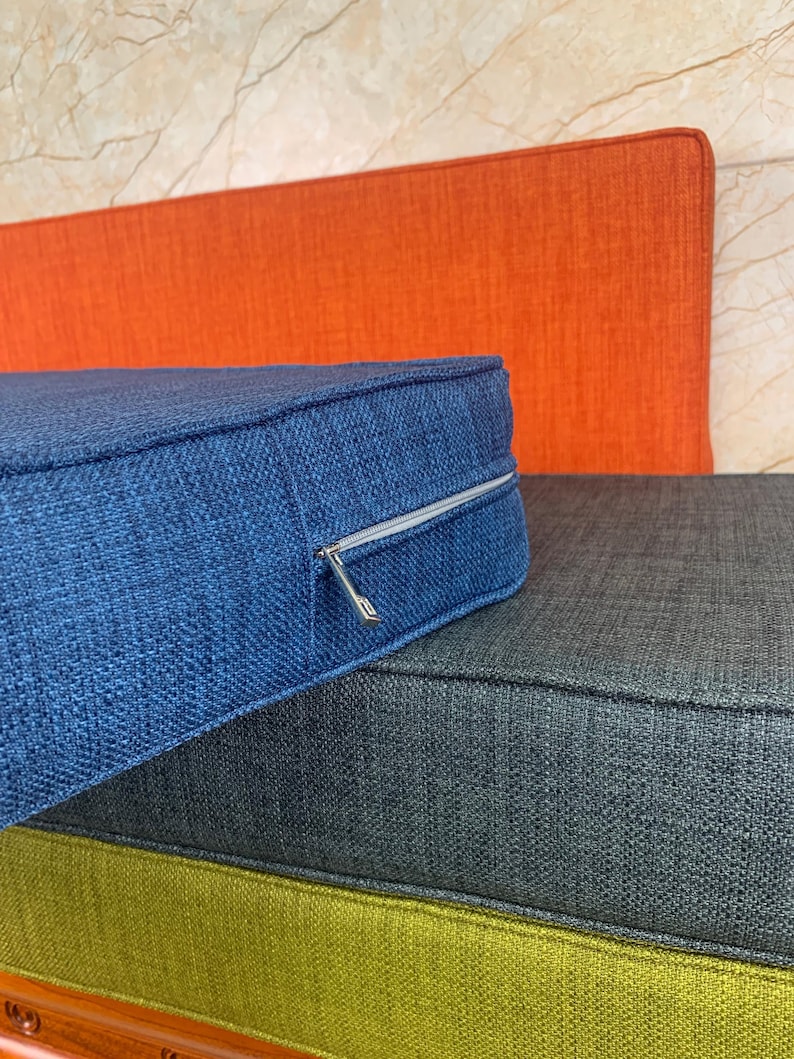 May include: Three cushions stacked on top of each other. The top cushion is blue, the middle cushion is gray, and the bottom cushion is green. The blue cushion has a zipper.