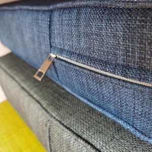 May include: A close-up of a blue fabric cushion with a zipper detail. The cushion is layered on top of two other cushions, one gray and one yellow.