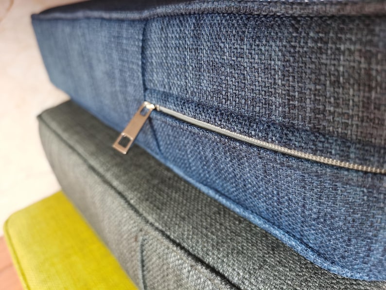 May include: A close-up of a blue fabric cushion with a zipper detail. The cushion is layered on top of two other cushions, one gray and one yellow.