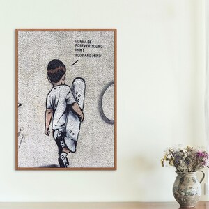 Pu&ograve; includere: Una stampa incorniciata di un murale di street art che presenta un giovane che si allontana dallo spettatore, portando uno skateboard. Il murale &egrave; su un muro bianco con il testo "Gonna be forever young in my body and mind" scritto su di esso.