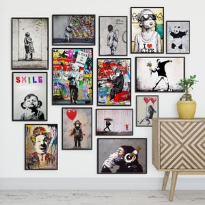 May include: A collection of framed art prints featuring various street art designs. The artwork includes images of children, monkeys, and other figures, with bold colors and graffiti-style backgrounds. The prints are displayed on a white wall.