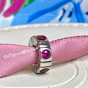 May include: A silver charm with three oval and three round magenta-colored stones. The charm is on a pink ribbon. The charm has the letters "ALE" and "D2G" engraved on it.