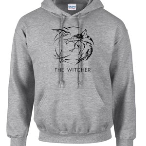 K&ouml;nnte beinhalten: Ein grauer Kapuzenpullover mit einem schwarzen Aufdruck eines Wolfskopfes und dem Text "THE WITCHER" auf der Vorderseite.