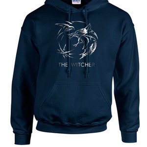 K&ouml;nnte beinhalten: Ein marineblauer Kapuzenpullover mit einem silbernen Wolf-Motiv und dem Schriftzug "THE WITCHER" auf der Vorderseite.