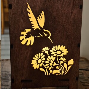 May include: A wooden lamp with a hummingbird and flowers cut out design. The lamp is lit and the cutouts are illuminated with a warm yellow light.