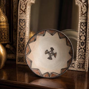 Peut inclure: Un bol en céramique peu profond avec un intérieur crème orné d'un symbole géométrique noir central et d'une bordure de motifs triangulaires hachurés noirs et terre cuite le long du bord intérieur avec un contour extérieur sombre.