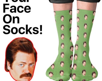 Peut inclure: Chaussettes vertes avec un motif répété d'un visage d'homme. Le texte "Your Face On Socks!" est affiché en noir. Les chaussettes sont portées par une personne.