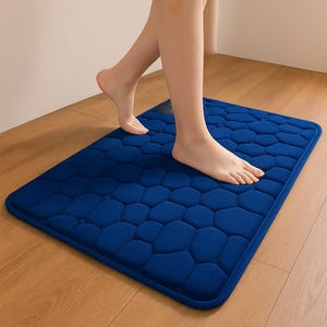 May include: A blue bath mat with a pebble-like design. The rectangular mat is on a wooden floor. The mat is approximately 24 inches long and 16 inches wide.