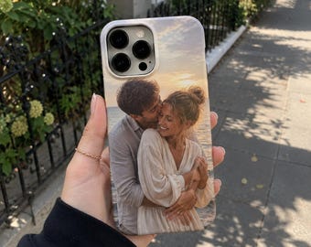 May include: A rectangular smartphone case featuring a printed photograph of two people embracing on a beach at sunset. The person on the left wears a light-colored shirt, while the person on the right wears a light-colored long-sleeved dress.